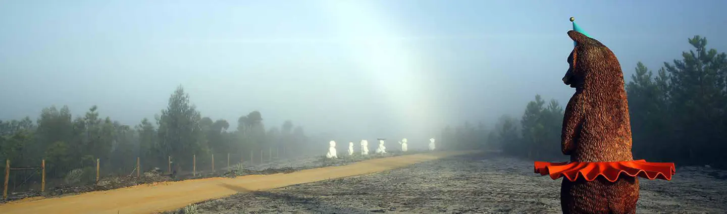 Foggy landscape with a wooden bear statue wearing a party hat and clown collar, and figures in white on a path.