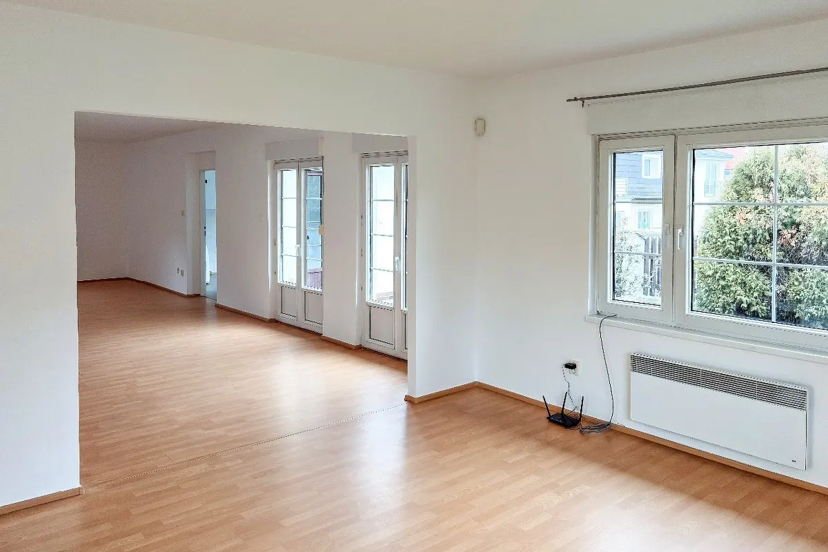 Bright, empty room with wood floors and white walls. A large window looks out onto greenery. French doors lead to another room.