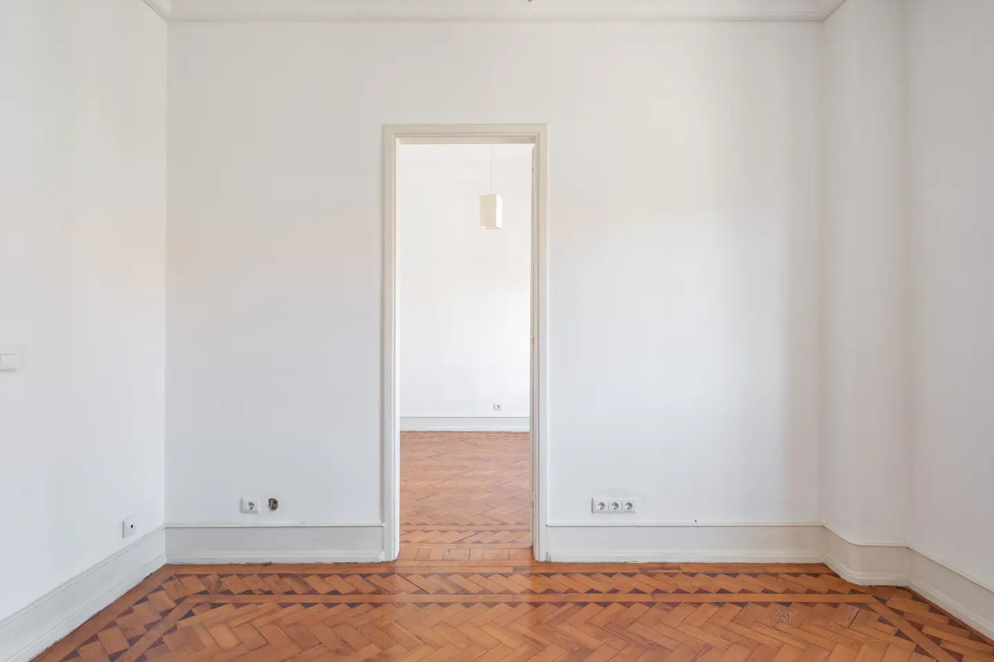Empty room with white walls, herringbone wood flooring, and a doorway leading to another bright, matching room.