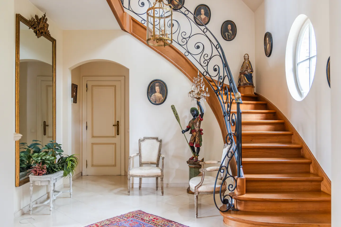 Elegant foyer with a curved wood staircase, black iron railing, and marble floors. Portraits adorn the walls. A large mirror and plants add to the ambiance.