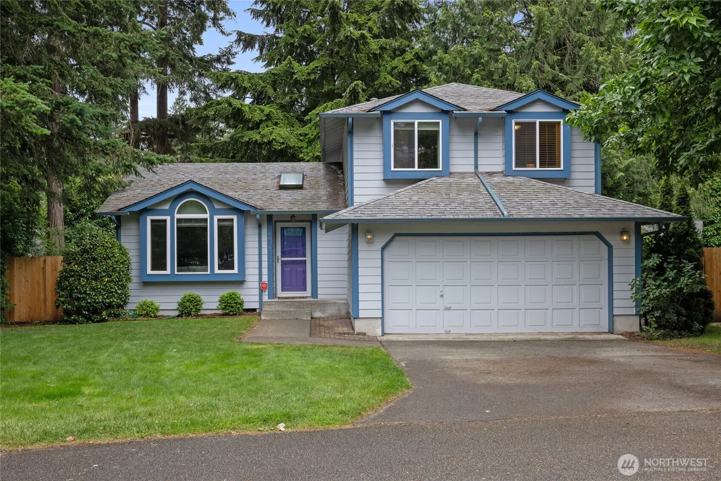 Two-story light blue house with a purple front door, white garage, and a green lawn surrounded by tall evergreen trees.