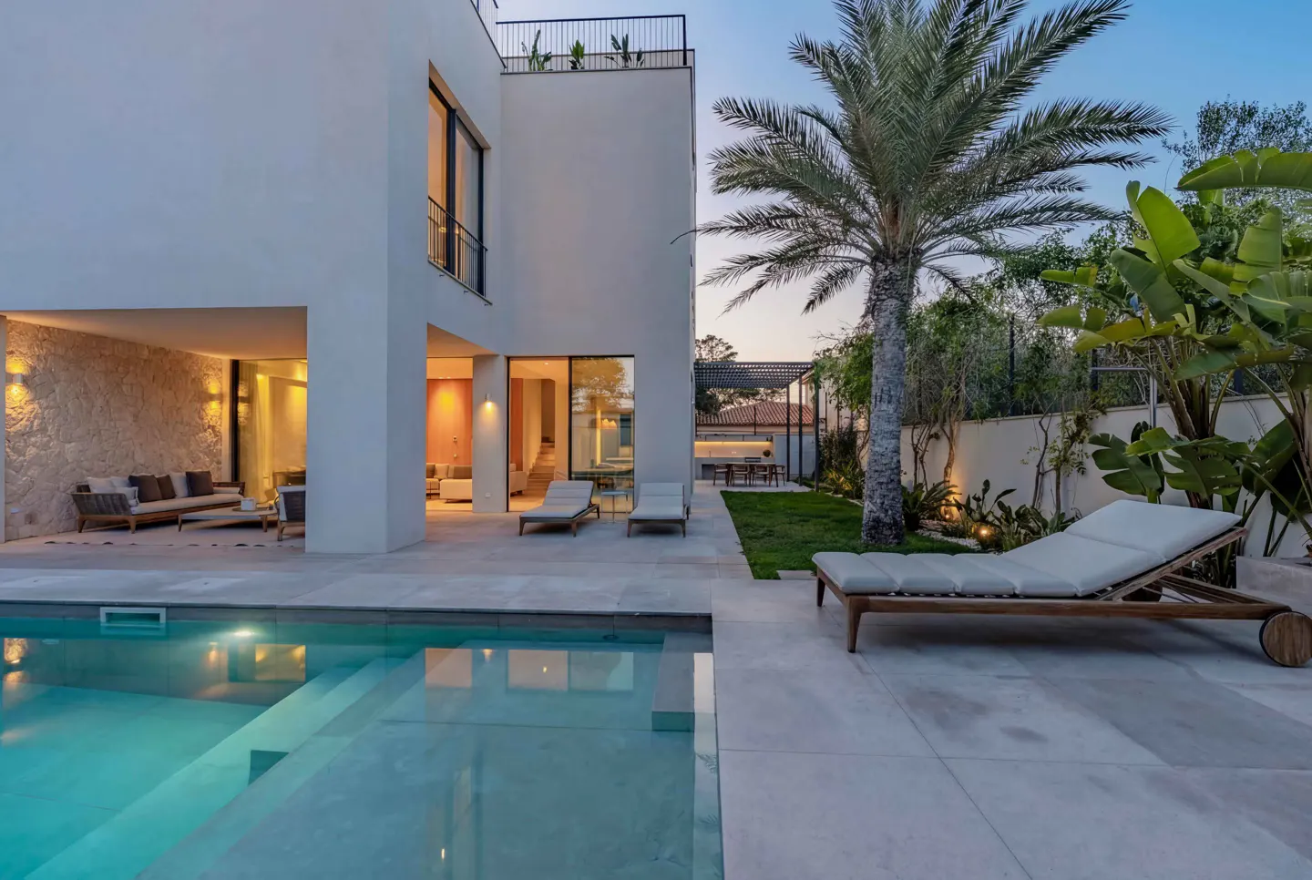 Modern white house exterior with a pool, lounge chairs, and palm tree at dusk.