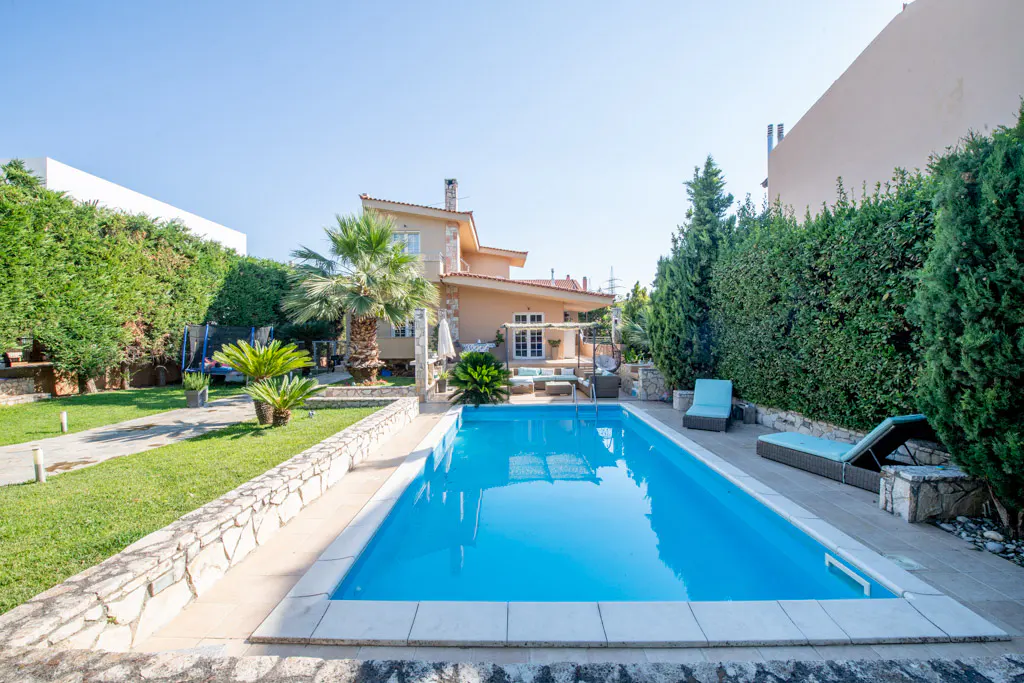 Backyard view of a rectangular blue swimming pool, lounge chairs, and a beige two-story house with a red tile roof.