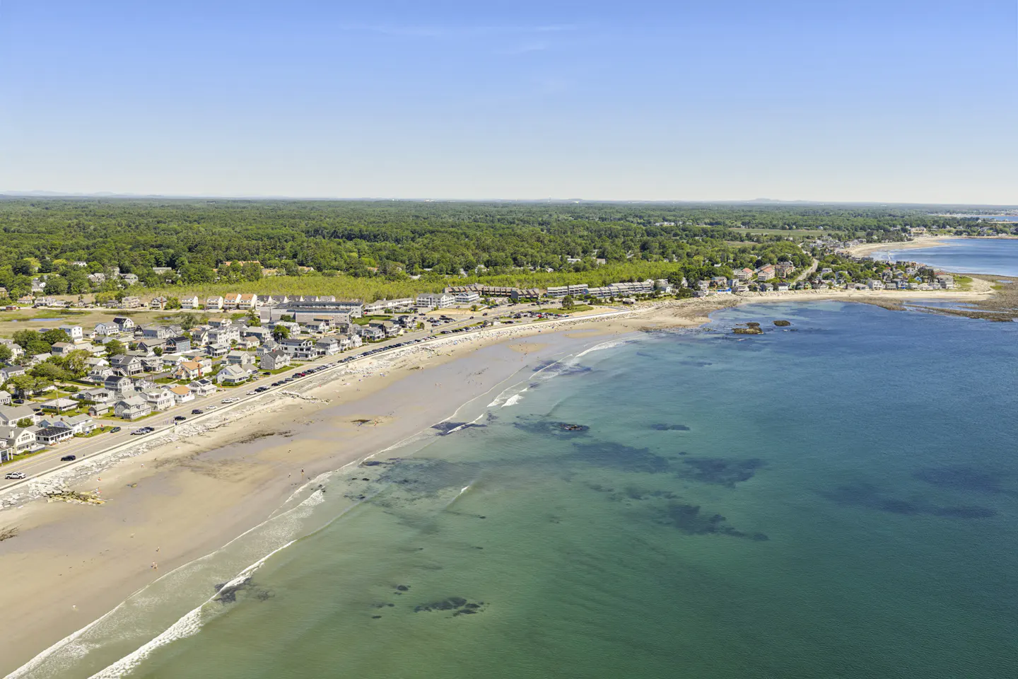 Aerial view of a sandy beach coastline with houses, a road, and dense green forest under a clear blue sky.