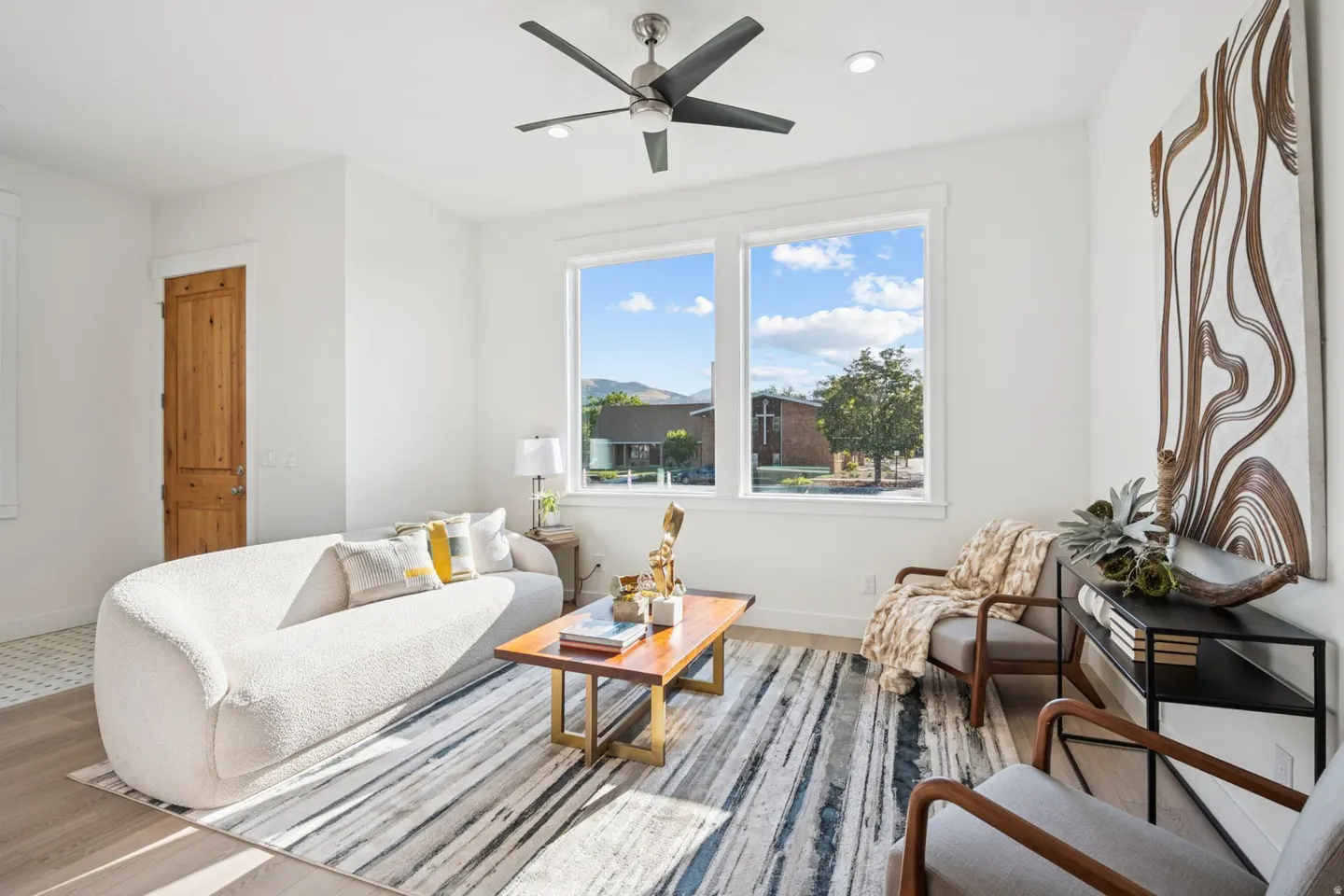 Bright living room with white walls, a curved white sofa, and a striped rug. A wooden coffee table sits in the center, and a large window offers a view of a building.