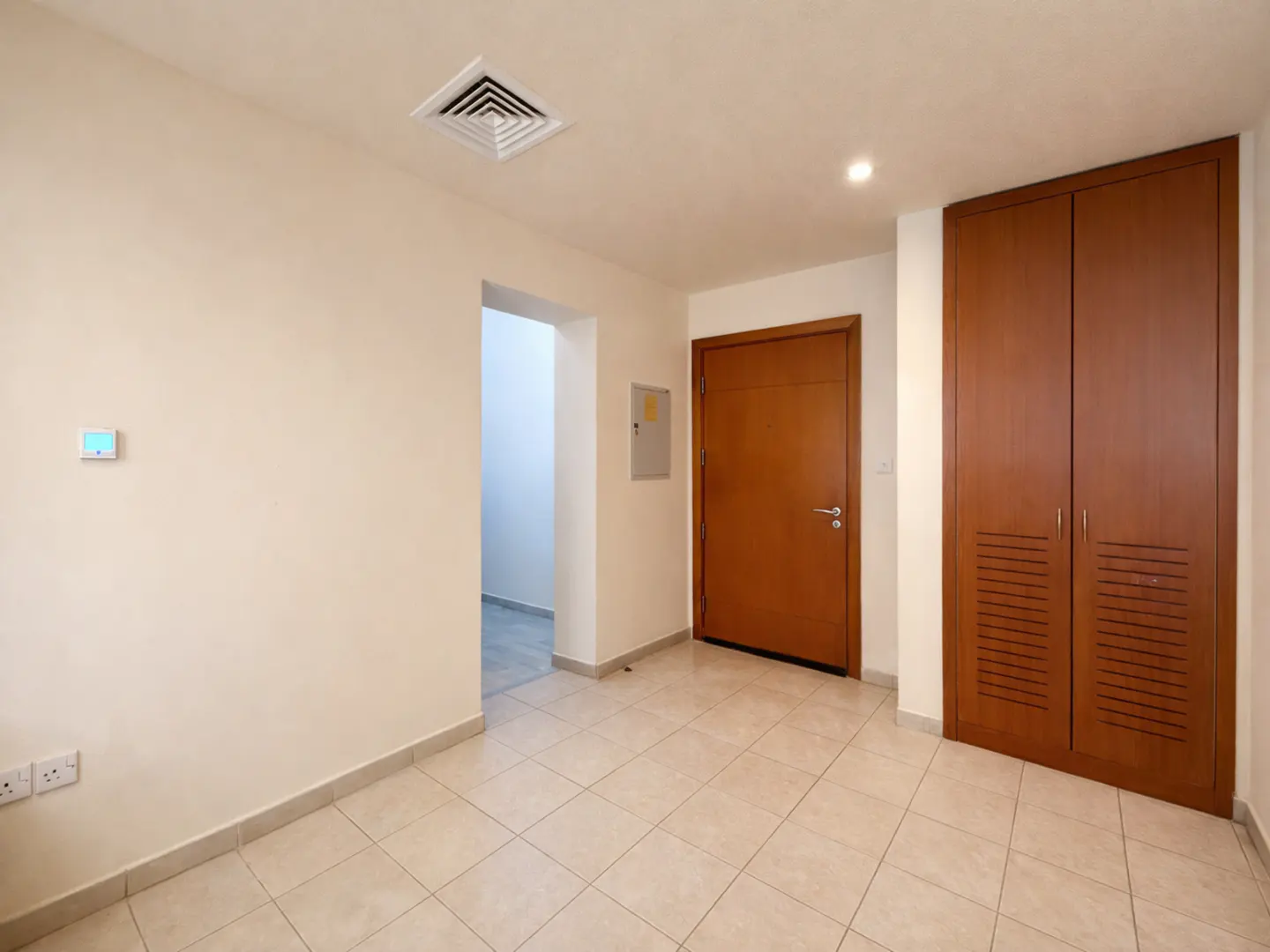 A bright room with beige tiled floors, a wooden door, a matching wooden closet, and an open doorway leading to a hallway.
