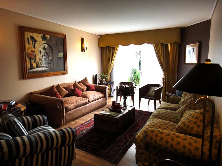 Living room with sofas, striped armchair, and trunk-style coffee table on a red rug. Curtains frame a bright doorway to a garden.