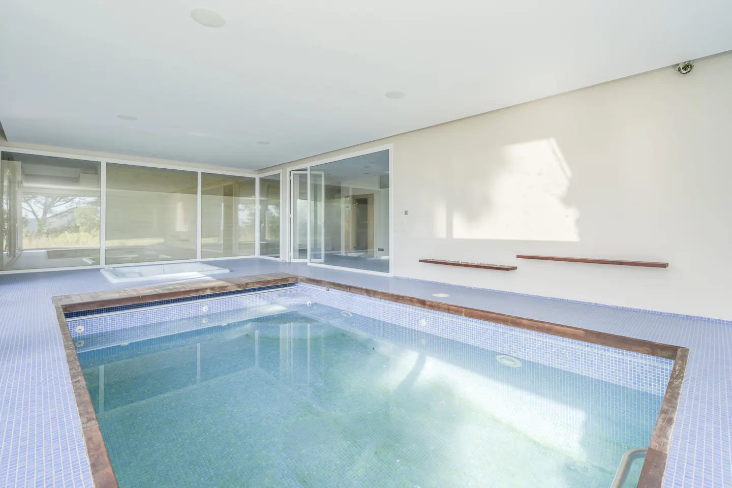 Indoor pool with blue tile and wood trim, surrounded by large windows and a white wall with floating shelves.