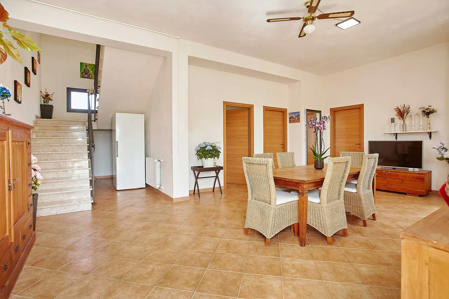 Bright, open dining room with tile floors, a wooden table with wicker chairs, and a staircase leading to the upper level.