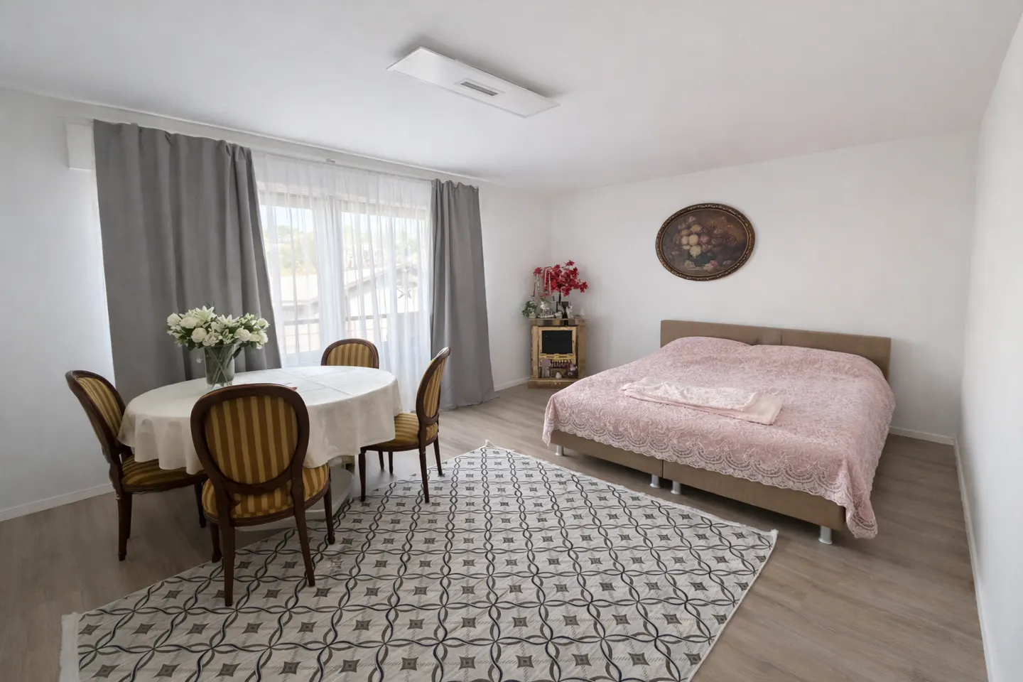 Bright studio apartment with a dining table, chairs, and a bed with pink bedding. Gray curtains frame a window.