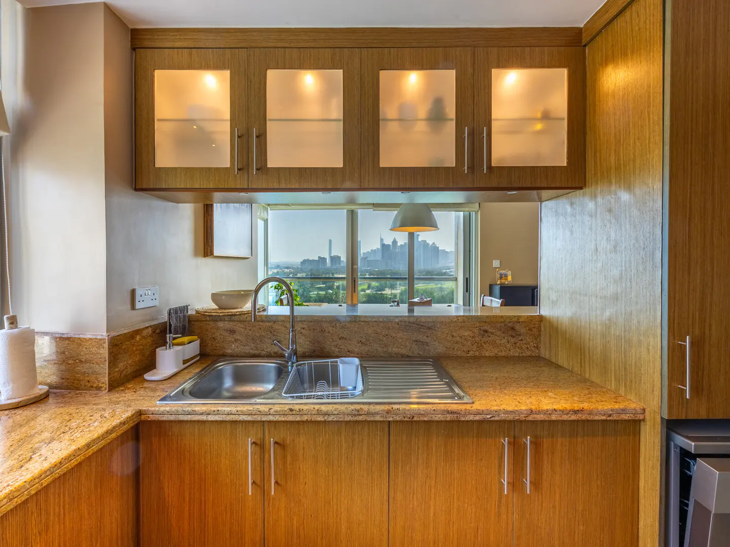 A kitchen with wood cabinets, granite countertops, and a stainless steel sink. A window overlooks a city skyline.