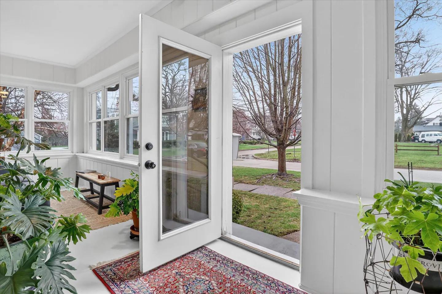 Sunroom with white walls, large windows, and an open glass door leading to a yard with a tree. Potted plants and a rug add color.