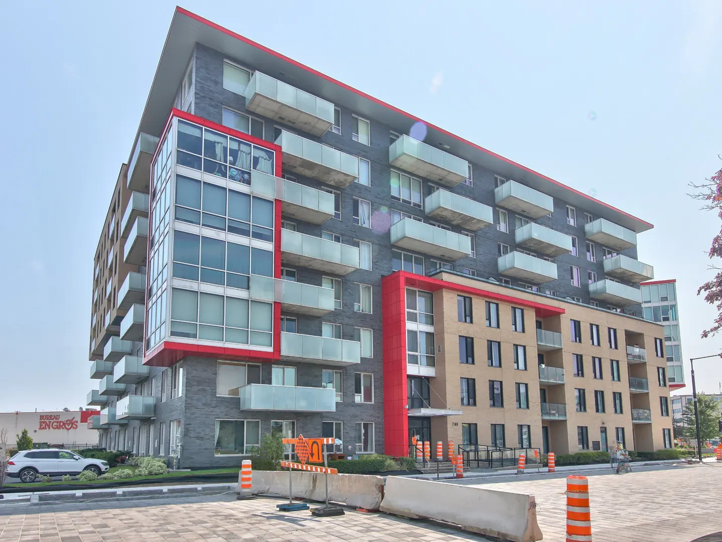 Modern multi-story apartment building with grey and tan brick, red accents, and glass balconies under a clear blue sky.