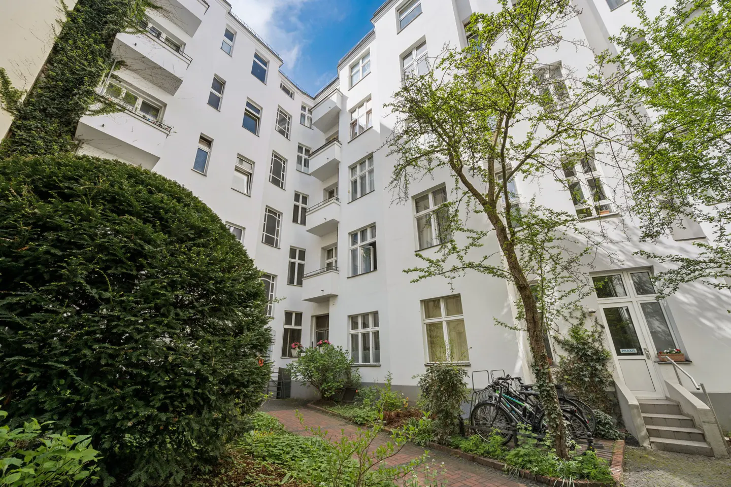 Courtyard view of a white apartment building with balconies, green trees, bushes, and parked bicycles.