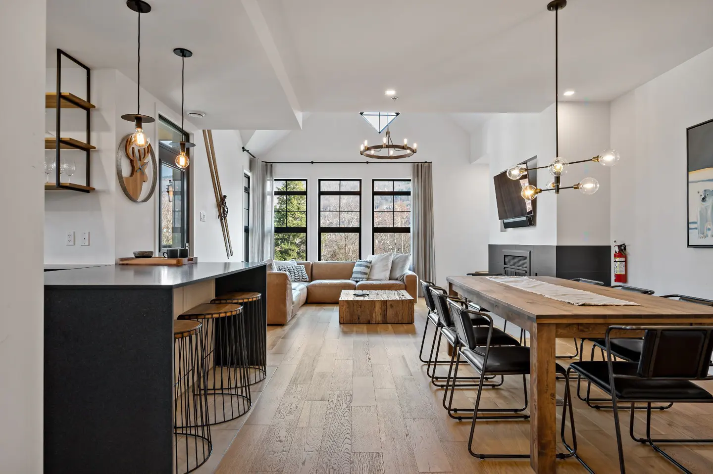 A modern living space with a kitchen island, a tan leather sofa, and a wooden dining table. Large windows offer a view of trees.