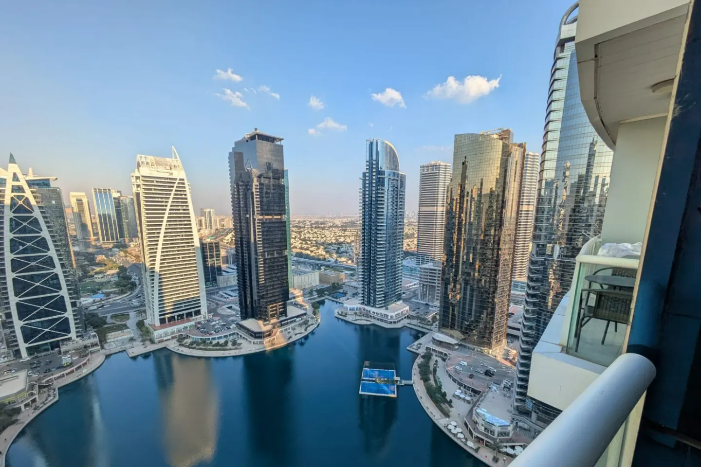 View from a high-rise balcony overlooking Dubai's skyline, featuring modern skyscrapers and a blue waterway. Balcony furniture is visible.