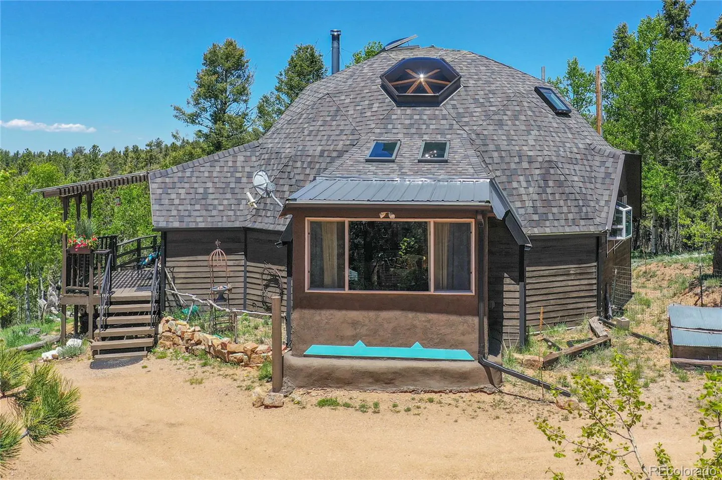 Exterior view of a dome-shaped house with a gray shingled roof, skylights, and a wooden deck surrounded by trees.