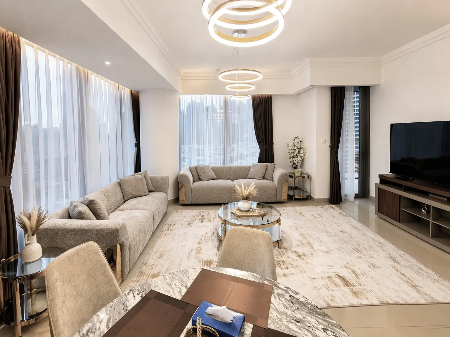 Bright living room with beige sofas, round glass table, and marble dining table. White walls, brown curtains, and modern ring chandeliers.