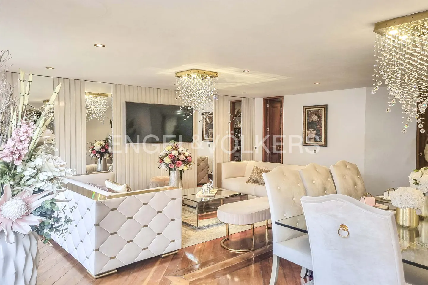 Living room with white sofas, glass table, wood floors, and crystal chandeliers. Flowers add color to the neutral palette.
