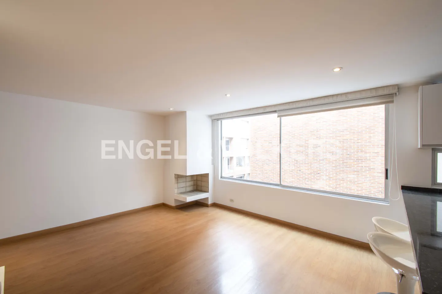 A bright, empty living room with hardwood floors, a fireplace, and a large window with a view of a brick building.
