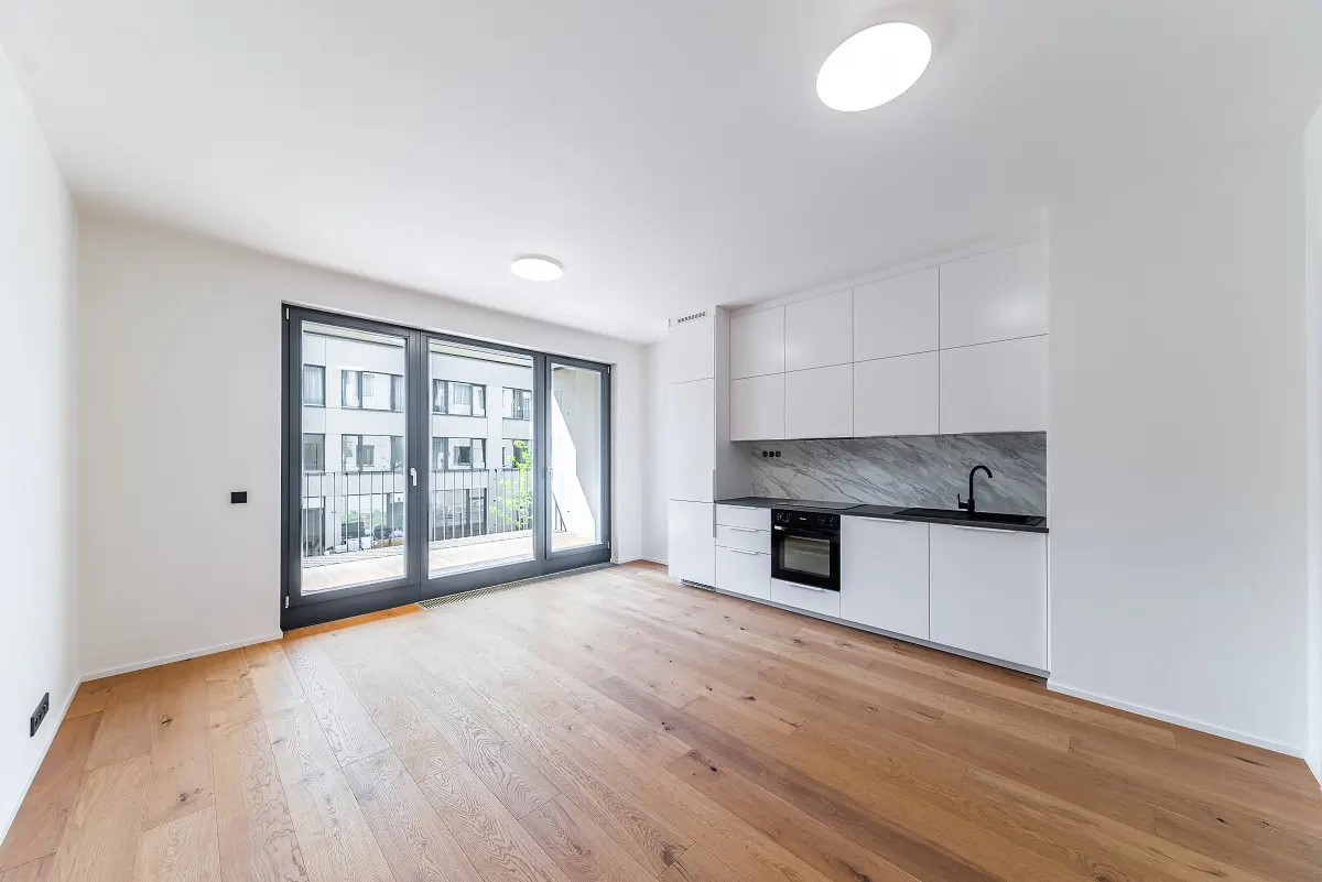 Bright, modern apartment with wood floors, white walls, and a kitchen with white cabinets and black countertops. Sliding glass doors lead to a balcony.