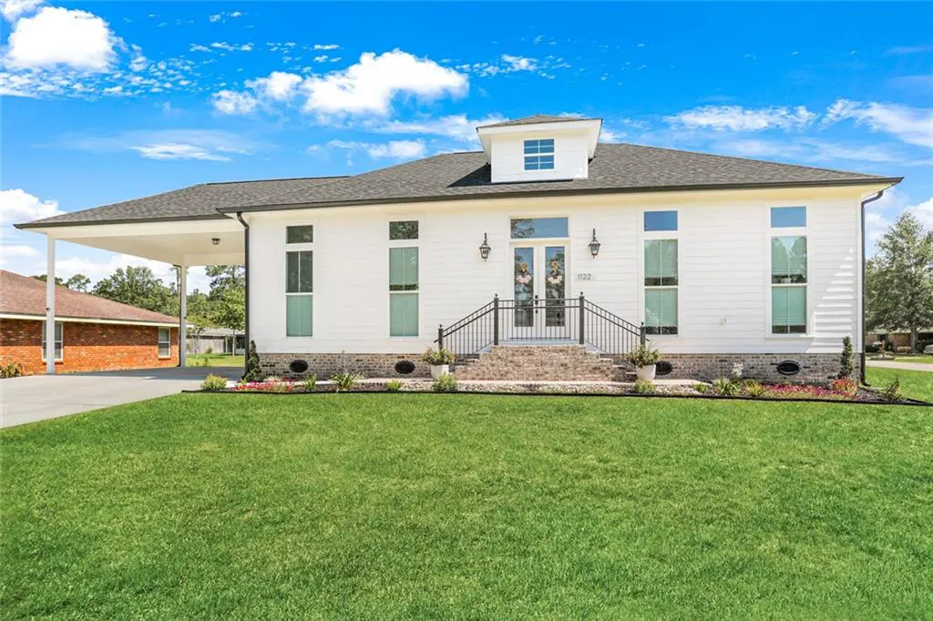 A white, one-story house with a black roof, a carport, and a green lawn under a blue sky. The house has a brick staircase with black railings.