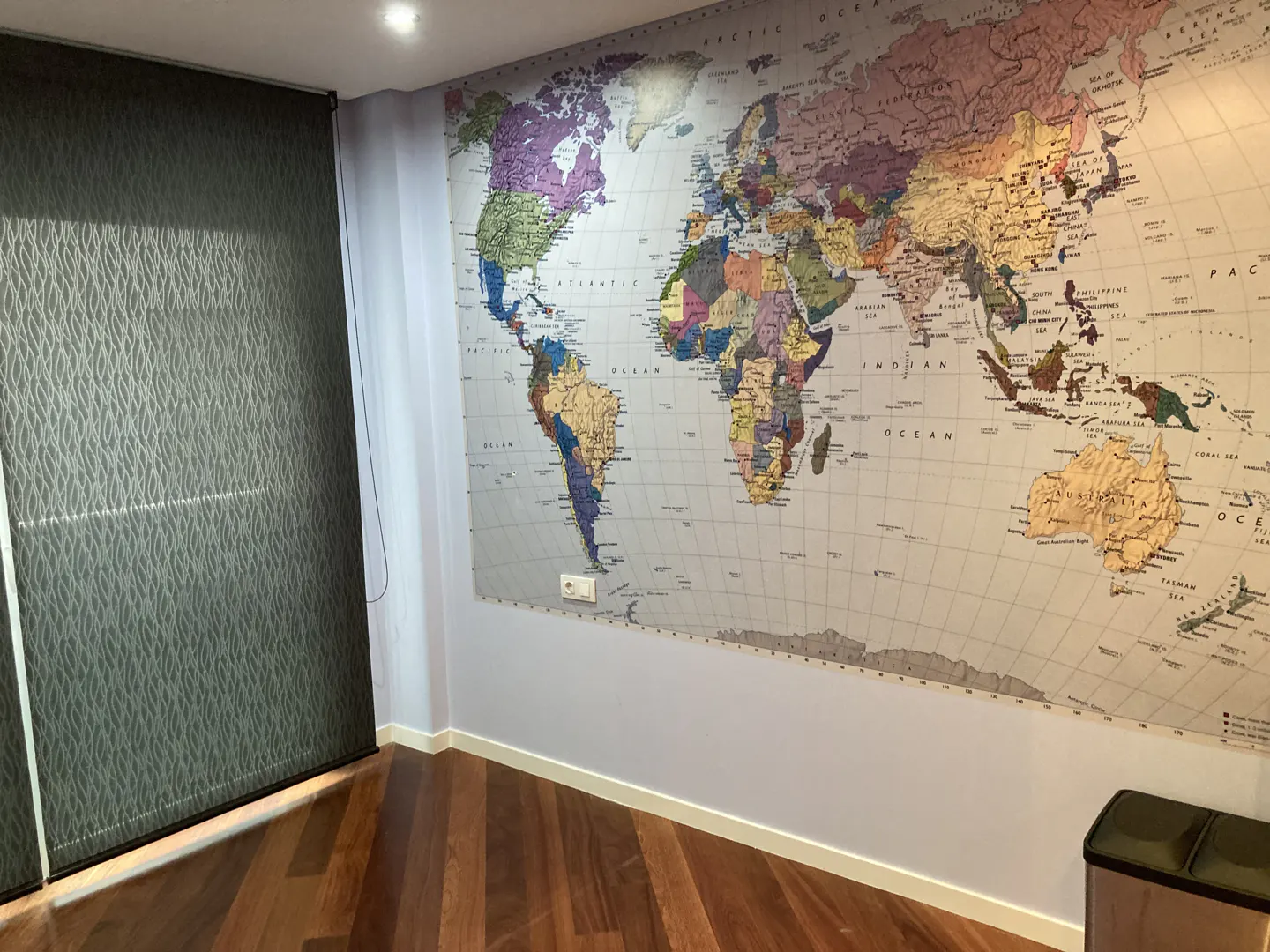 A room with a large world map on the wall, a dark window shade, and a wood floor.