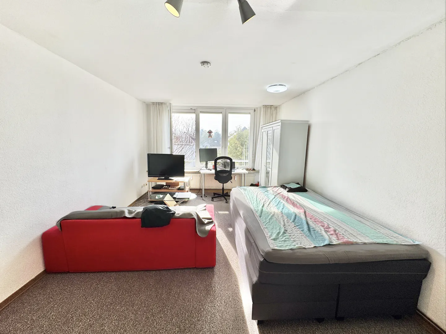 Bright studio apartment with a red sofa, bed, desk, and TV. White walls and carpeted floor. Natural light from a large window.