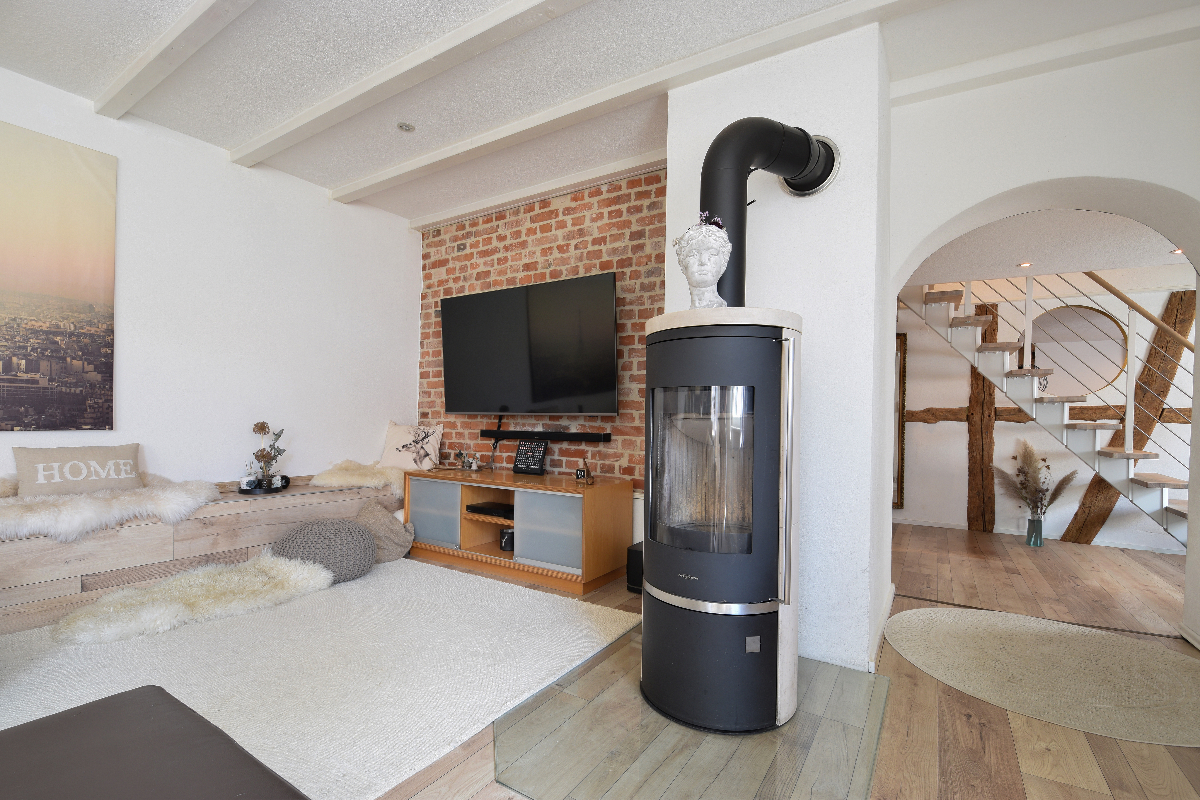 Modern living room with a brick accent wall, flat-screen TV, wood-burning stove, and an arched doorway leading to stairs. [AI-generated text]