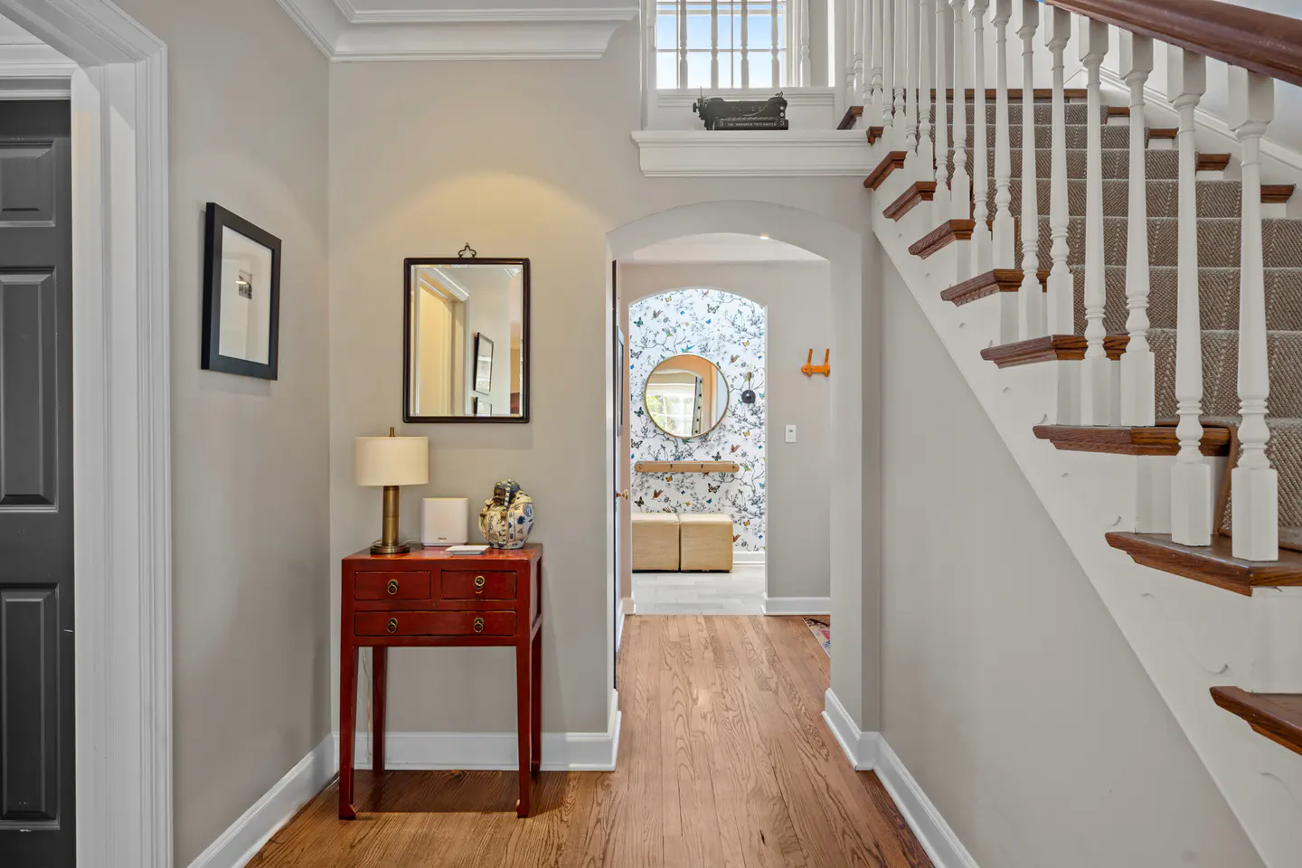 Foyer with hardwood floors, red accent table, and staircase. A mirror reflects a room with butterfly wallpaper.