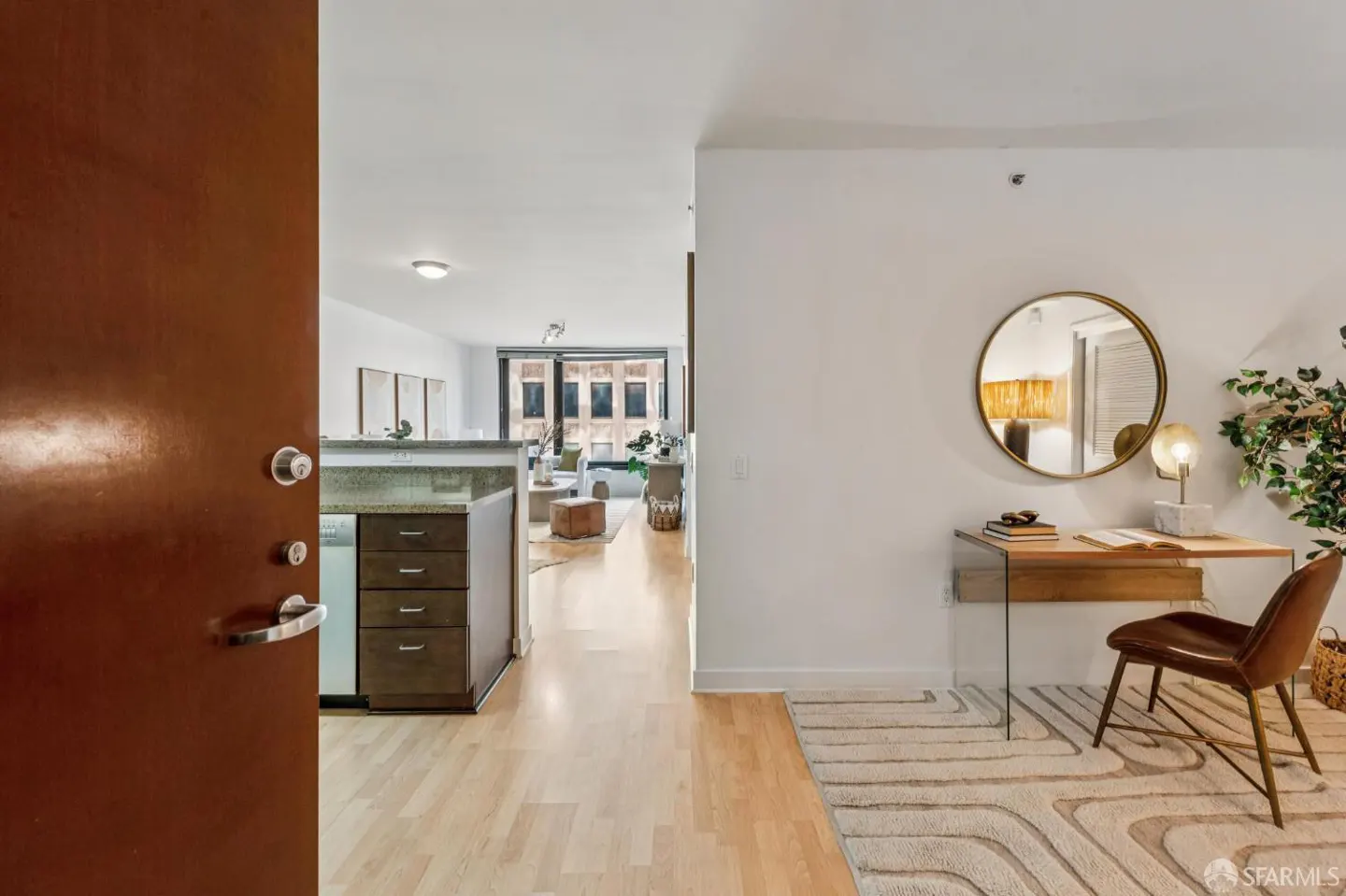 View from an open door into a modern apartment with a kitchen island, a desk area with a round mirror, and wood floors.