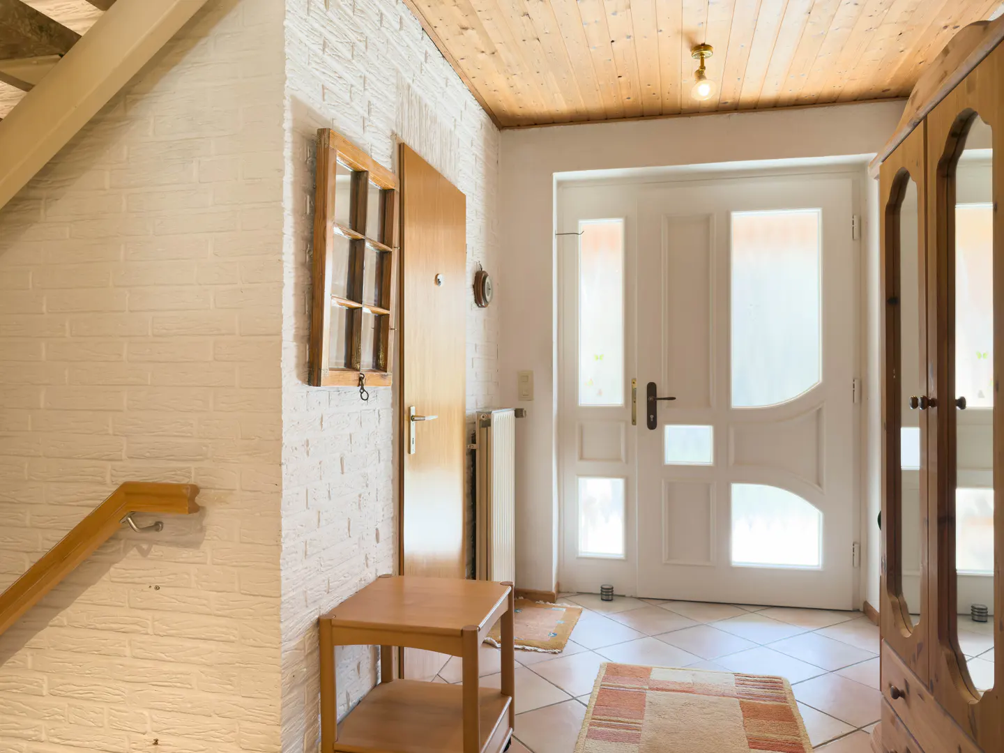 Entryway with white brick wall, wooden ceiling, and tiled floor. A wooden door and a white door are visible. A small wooden table sits near the wall.