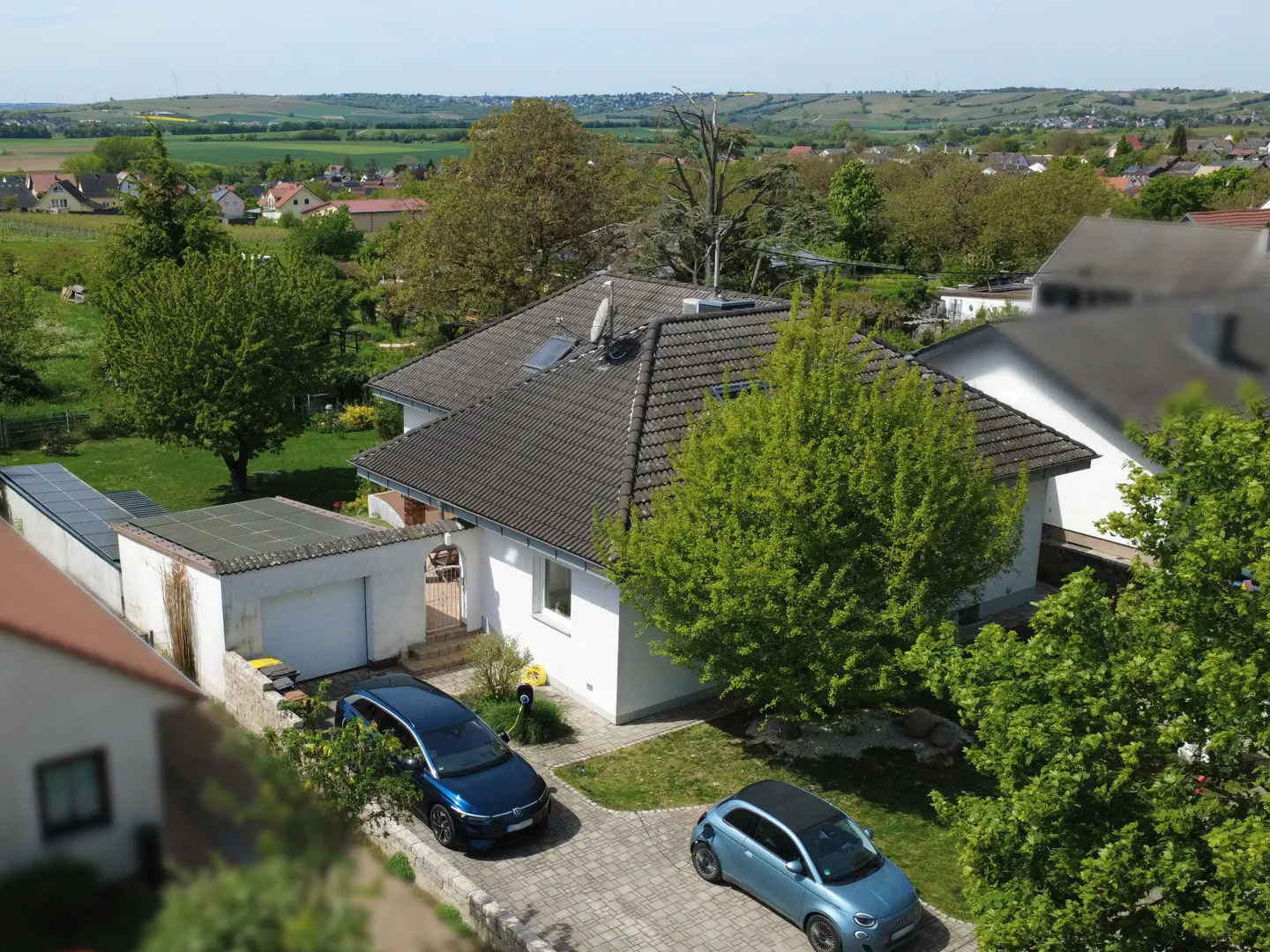 Aerial view of a white house with a gray roof, two cars parked in the driveway, and green trees in the background.