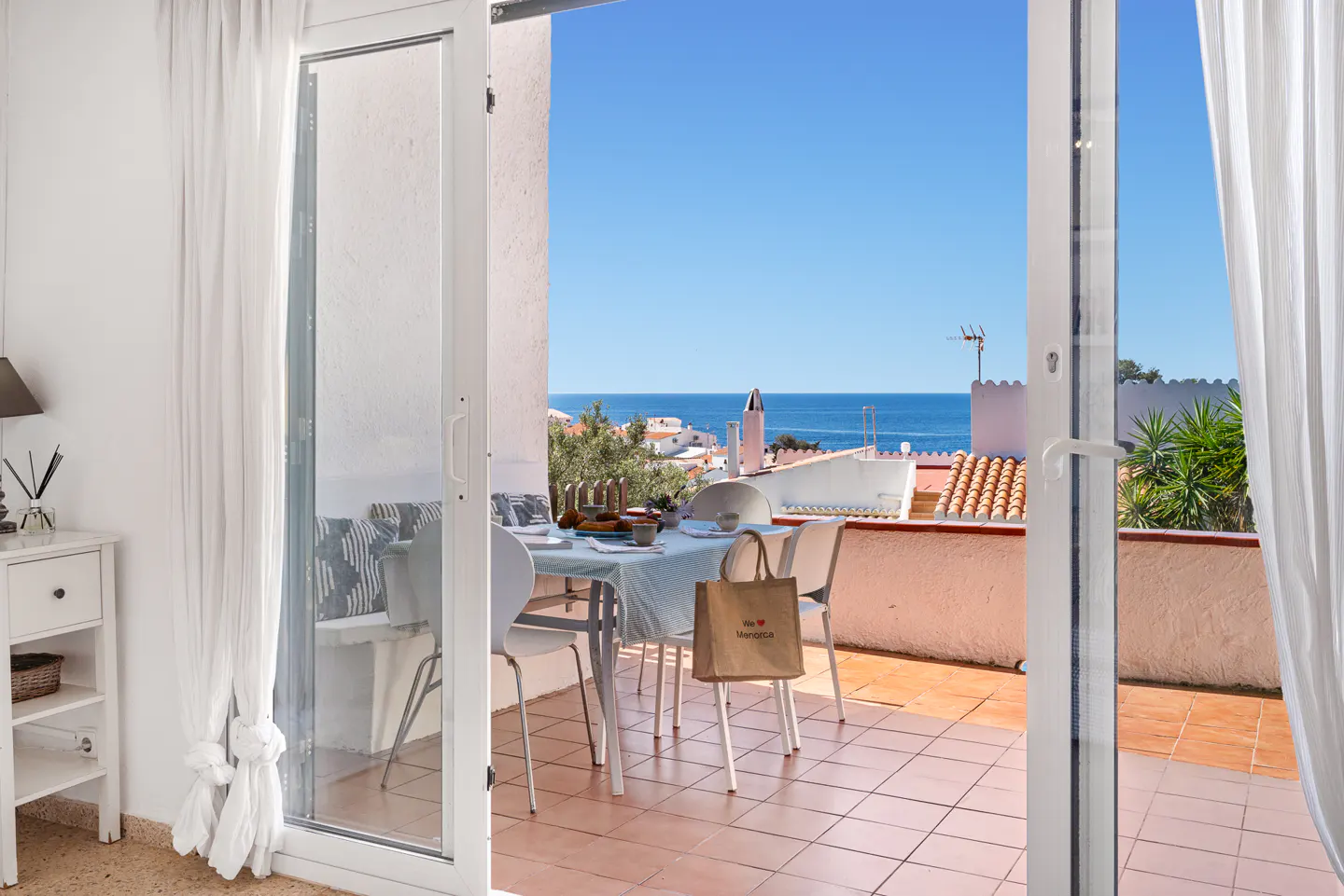Open patio doors reveal a table set for a meal, with an ocean view. A shopping bag rests on a chair.