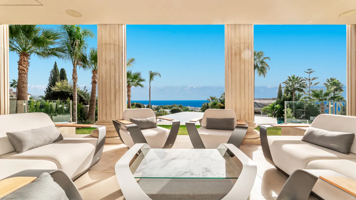 Luxury patio with white sofas, chairs, and a glass table. Tall columns frame a view of palm trees, the ocean, and a clear blue sky.