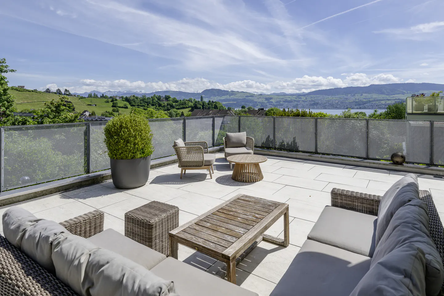 Outdoor patio with wicker furniture, a wooden table, and a potted plant. A metal fence surrounds the patio, with a view of green hills and a lake in the background.