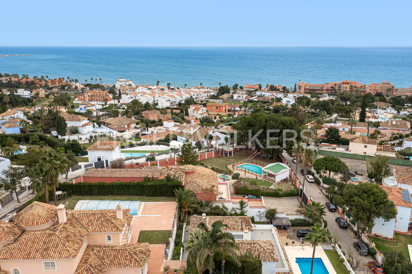 Aerial view of a coastal town with red-tiled roofs, green trees, and the blue ocean in the background.