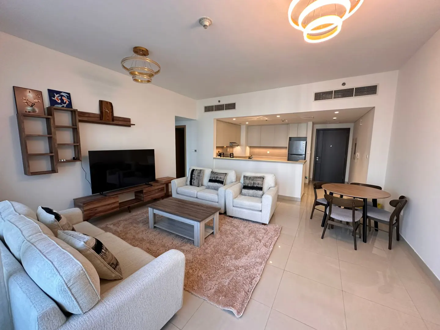 Bright, modern living room with white sofas, wood furniture, and a pink rug. A kitchen is visible in the background.