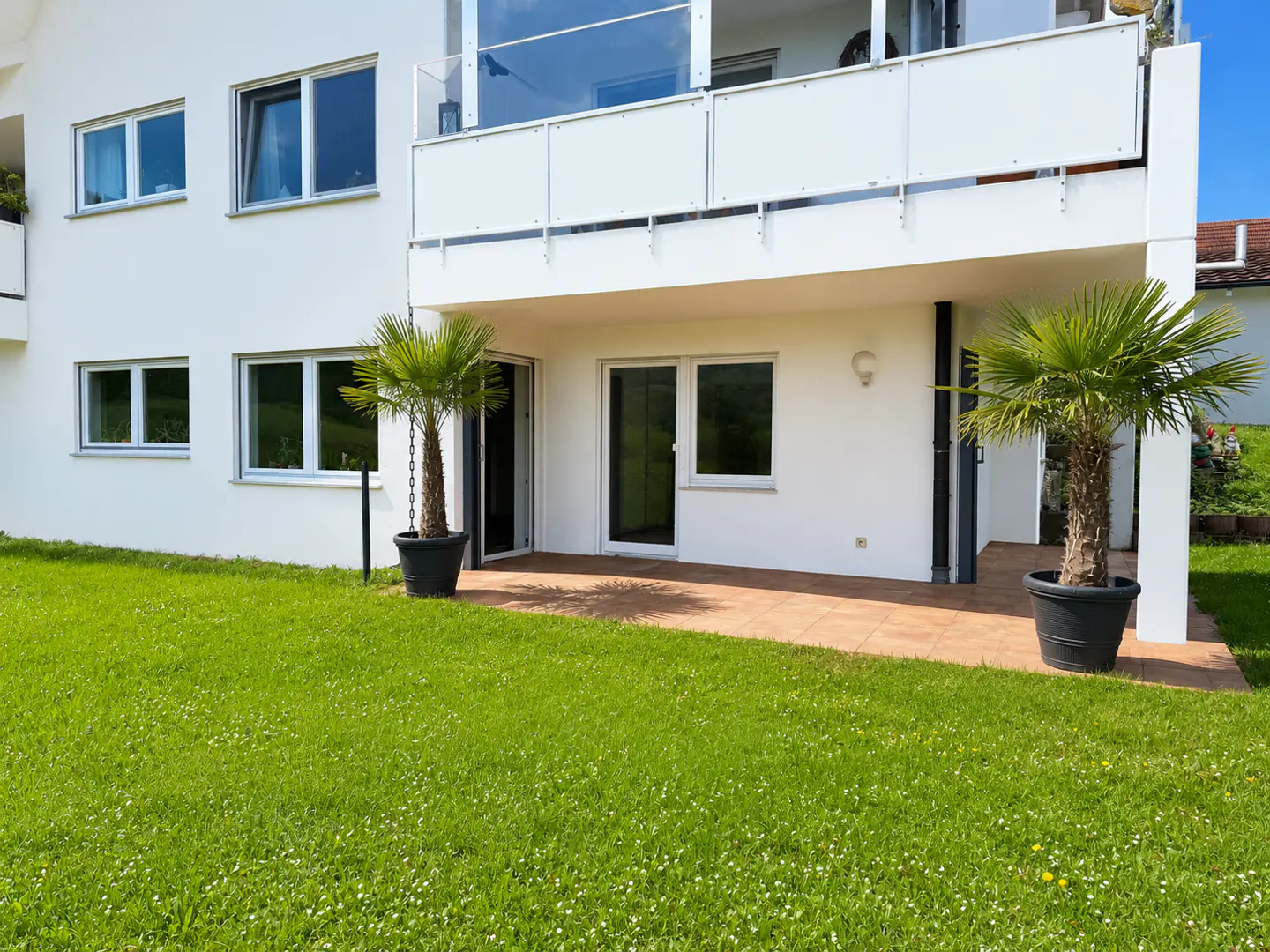 Exterior of a white two-story building with a balcony, patio, and green lawn. Palm trees in pots flank the patio doors.