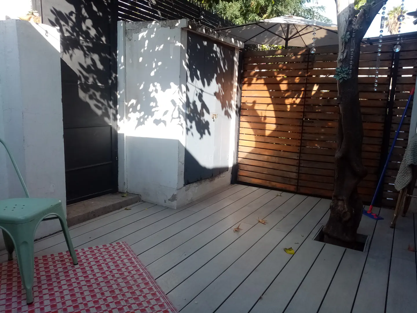 Outdoor patio with white wood flooring, a green chair, and a red and white checkered rug. A wooden fence and a tree are in the background.
