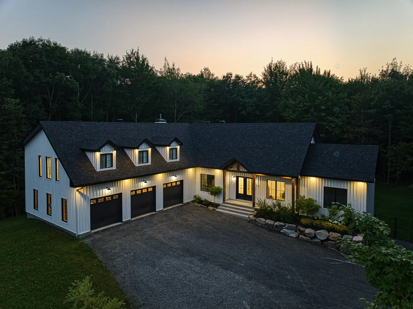 Exterior shot of a white farmhouse with a black roof, three-car garage, and gravel driveway at dusk.