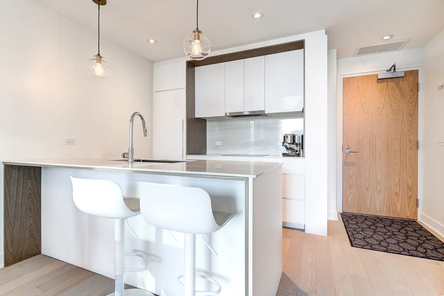 Modern kitchen with white cabinets, a breakfast bar with two white stools, pendant lighting, and a wooden entry door.