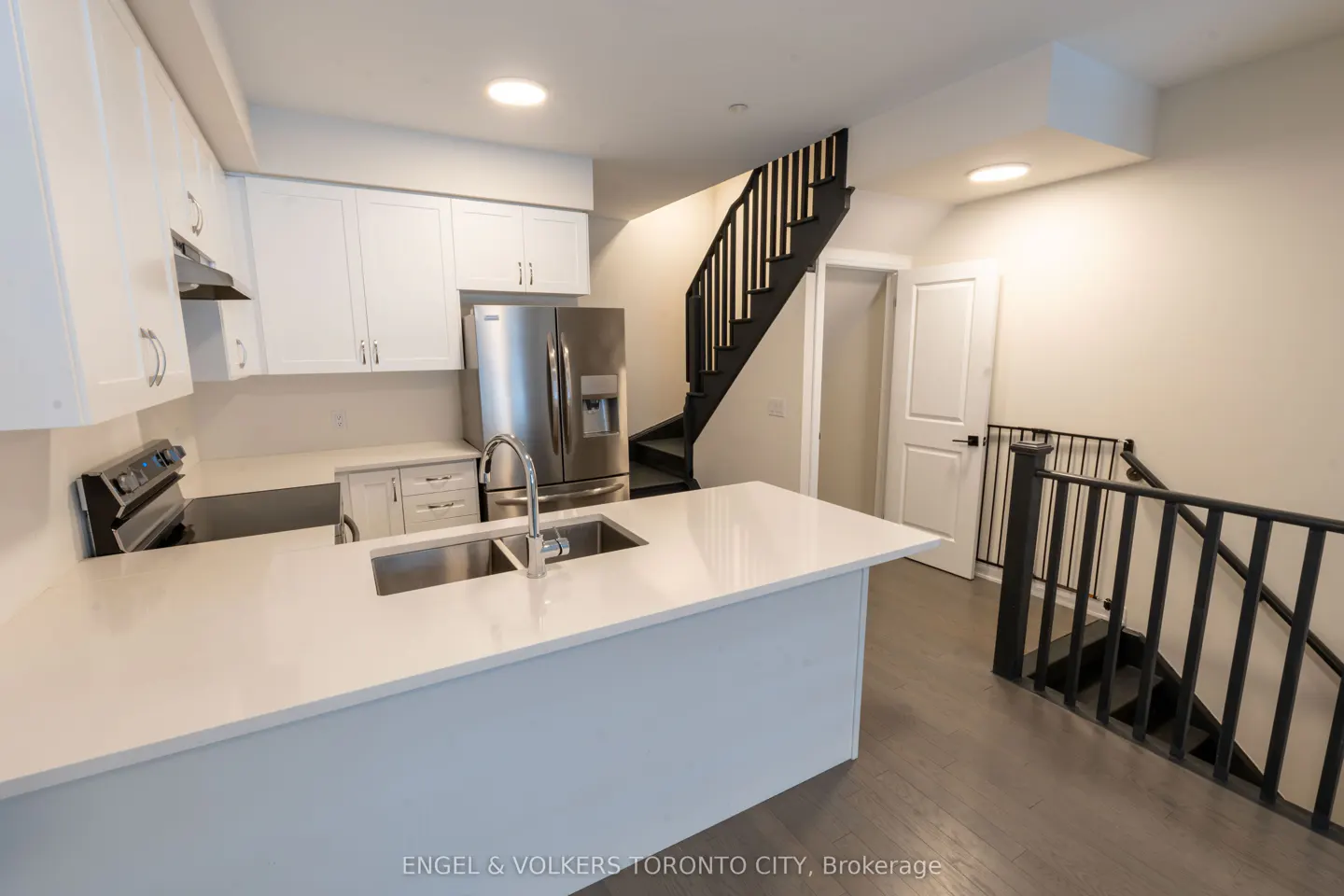 Modern kitchen with white cabinets, quartz island, stainless steel appliances, and a black staircase leading to a lower level.