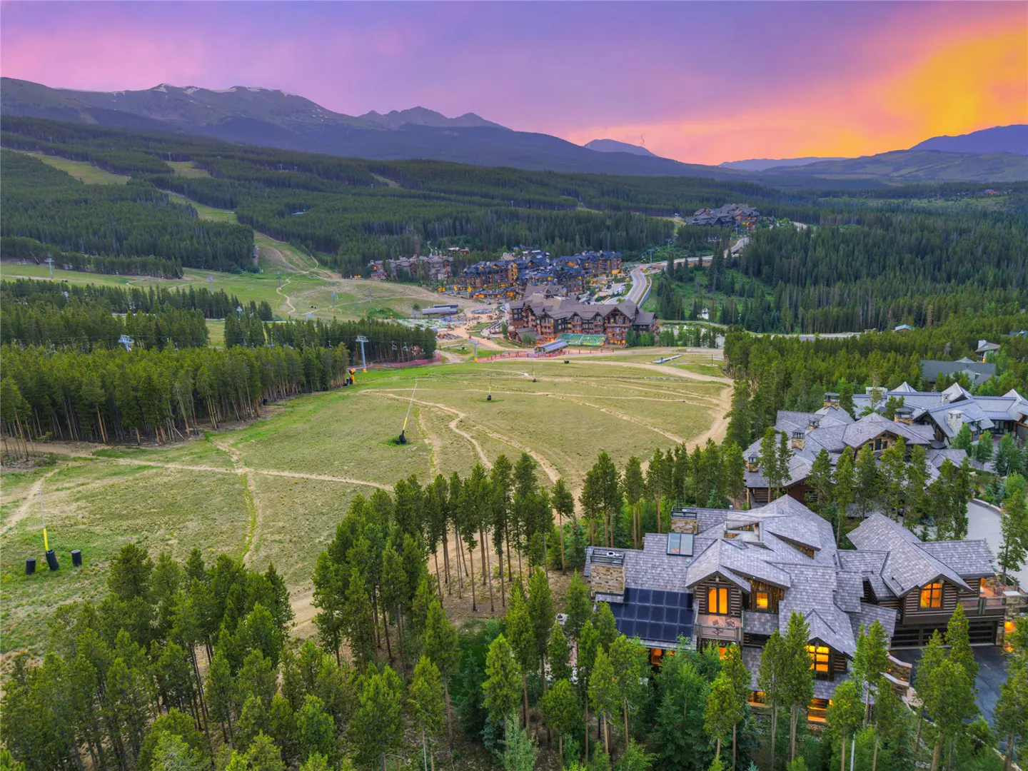 Aerial view of a luxury log home nestled in a forest, with mountains and a colorful sunset in the background.