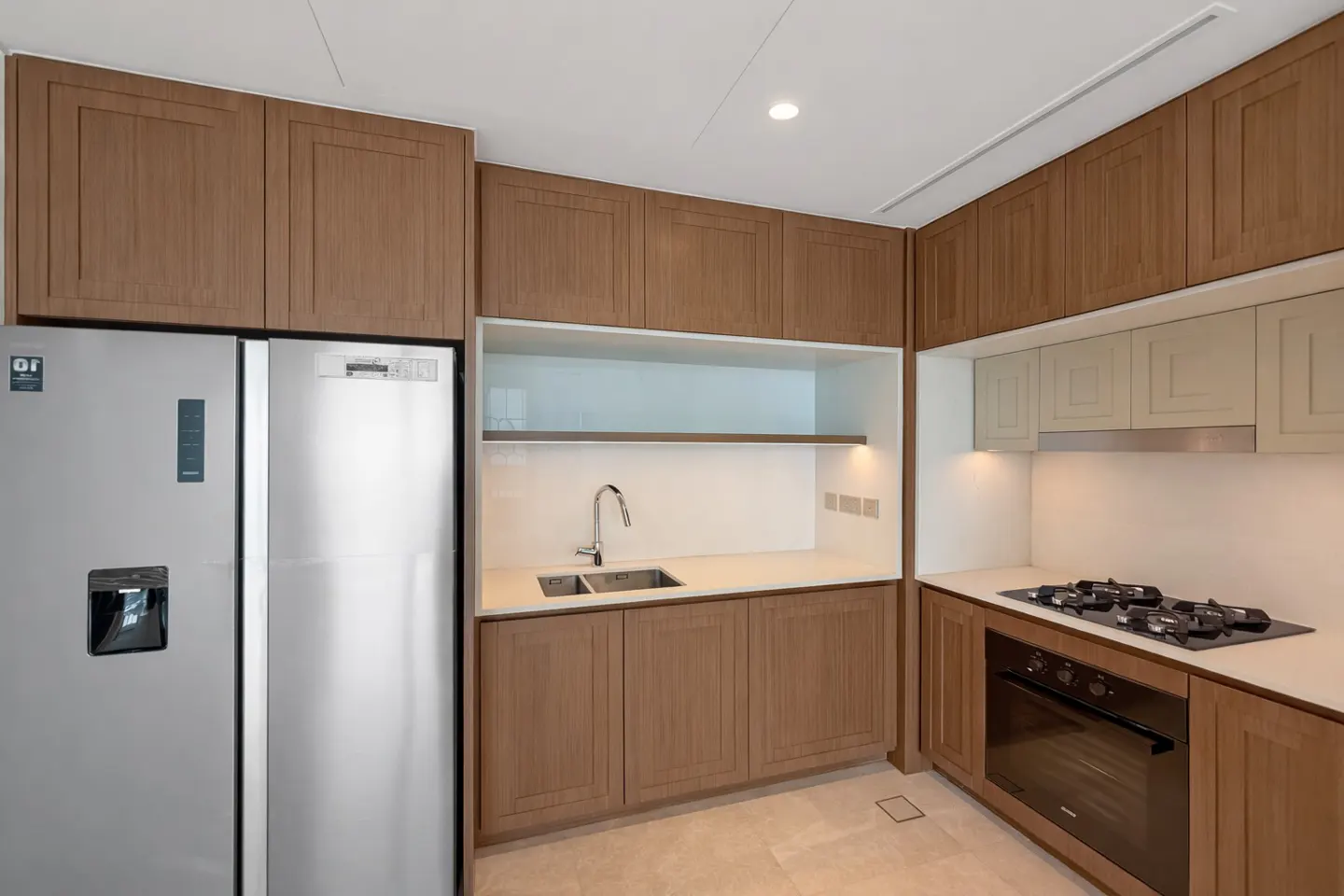 A modern kitchen with stainless steel refrigerator, wood cabinets, white countertops, and a gas stovetop.