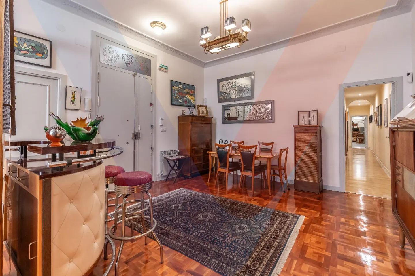 A living room with a bar, dining table, and hallway. The room has wood floors, a blue rug, and white walls with art.
