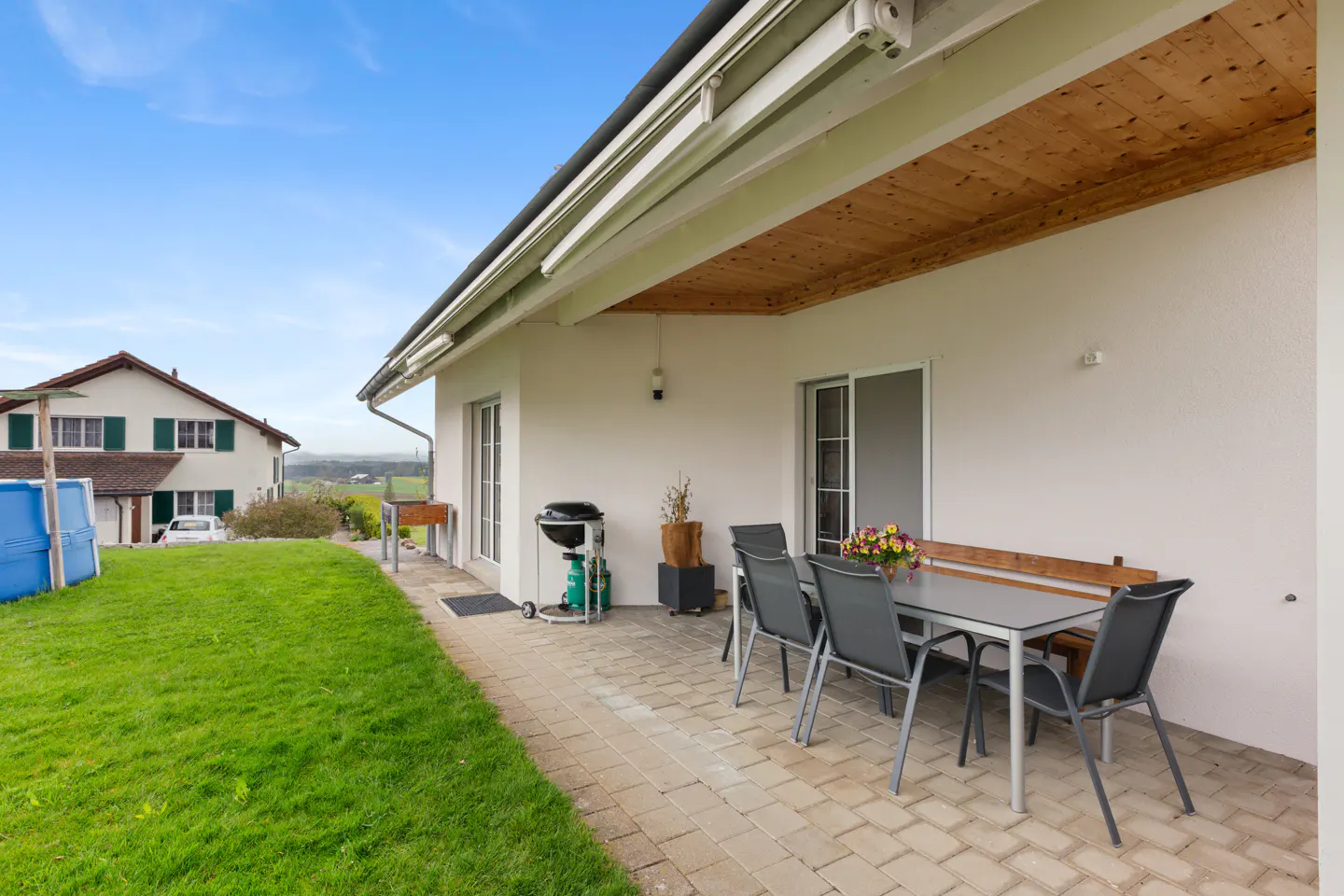 Covered patio with table, chairs, and grill. A lawn and house are in the background.