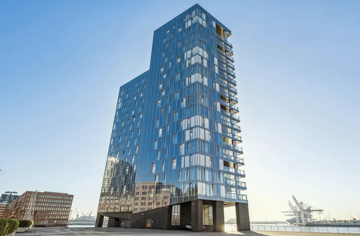 A tall, modern glass apartment tower with balconies stands against a clear blue sky near a harbor with industrial cranes.