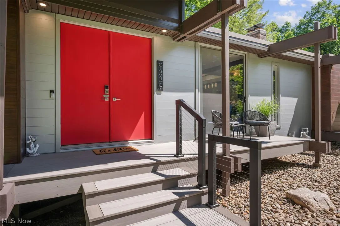 Modern home entrance with bright red double doors, gray wooden deck, cable railing, and two patio chairs.