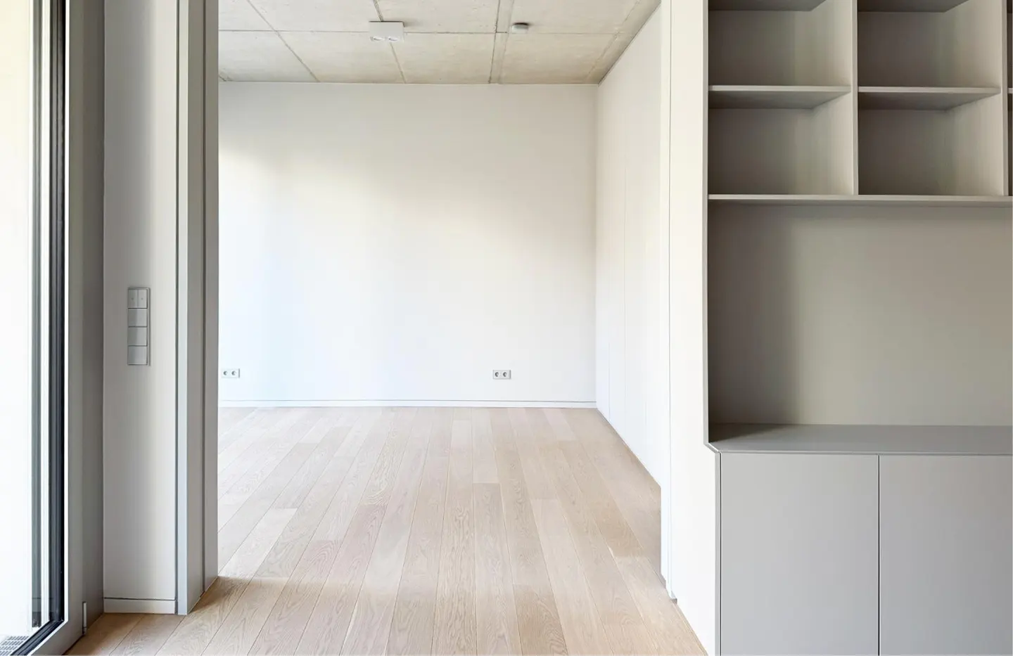 An interior view of an empty room with light wood floors, white walls, and a gray built-in shelving unit.